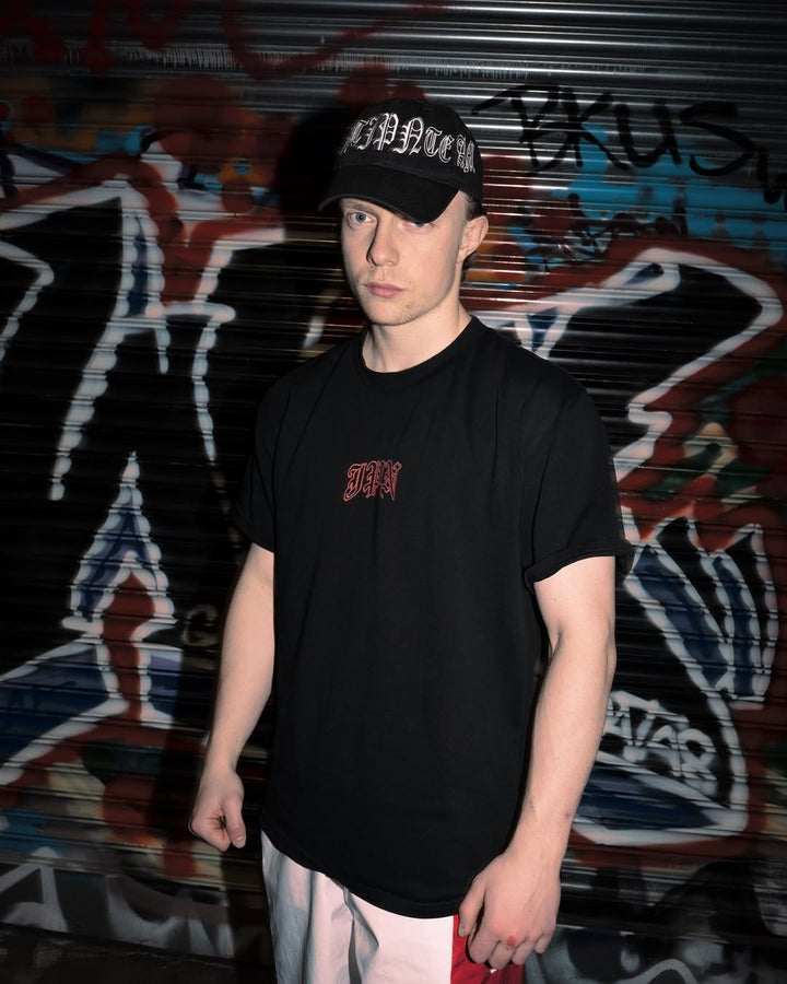 A man in the black JPN Hannya Tee and a black cap standing in front of a graffiti-covered shutter