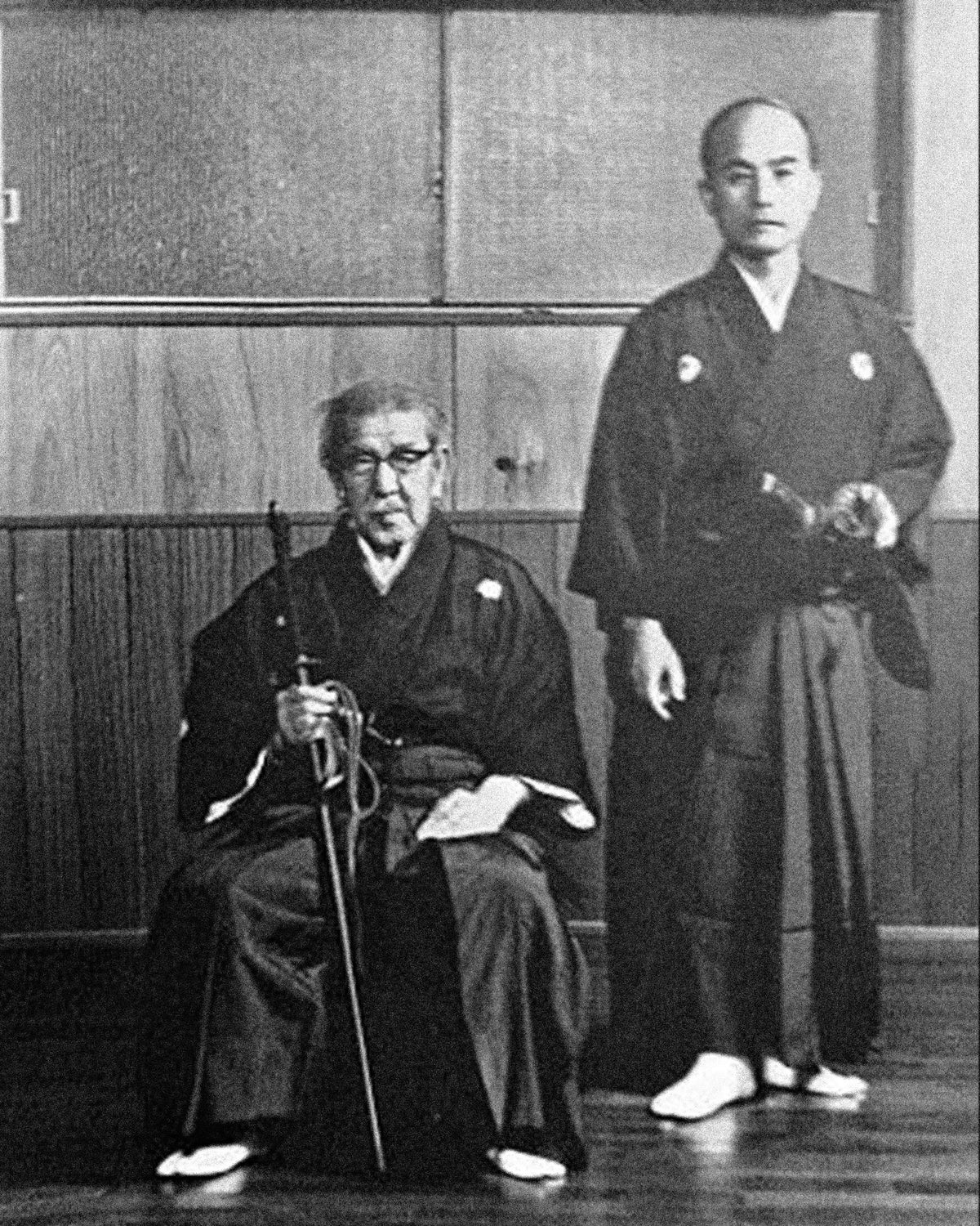 A sword master seated in a dojo with his student standing beside him